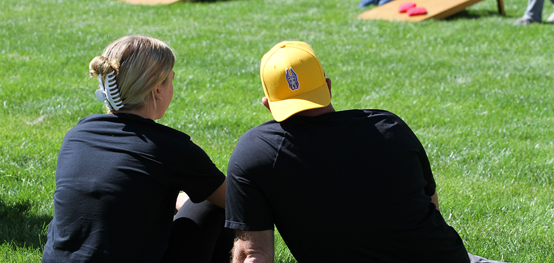 Two people sit on a lawn of grass during Family Weekend, watching the goings on around them.