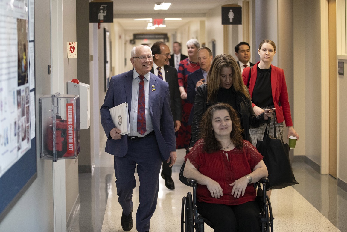 Congressman Paul Tonko tours the RNA Institute alongside members of UAlbany leadership. In this image, nine people are proceeding down a hall in the RNA Institute, among the labs. The group is led by Paul Tonko on the left and guest speaker Jennifer Rittner is being pushed in a wheelchair on the right. UAlbany President Havidán Rodríguez and RNA Institute Director Andy Berglund are in conversation behind them.