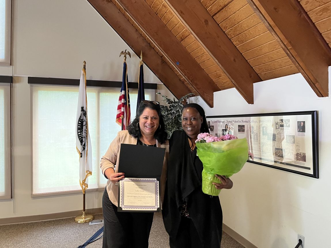 Two women stand next to each other, one holding a certificate, the other holding a pot of pale pink flowers wrapped in lime green paper. Both women are smiling. Three flags on vertical stands and a framed historical timeline are visible in the background. 