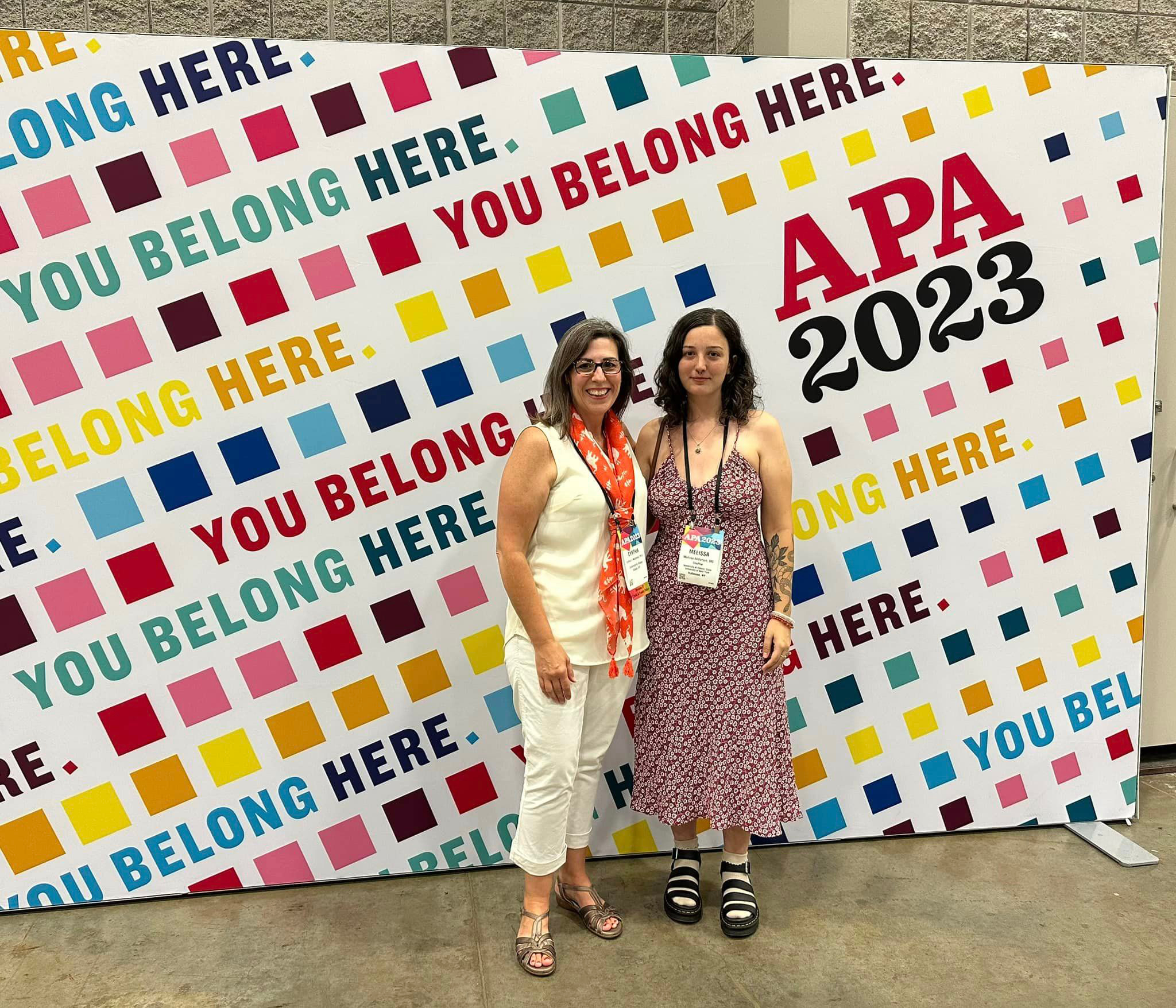 Two women wearing lanyards stand in front of a colorful pop-up wall that reads, 'you belong here.' and 'APA 2023'