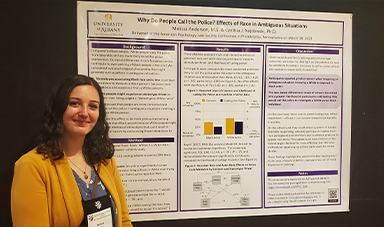 A woman with curly dark hair in a blue floral top and yellow blazer stands to the left of a research poster titled, 'Why Do People Call the Police? Effects of Race in Ambiguous Situations.'