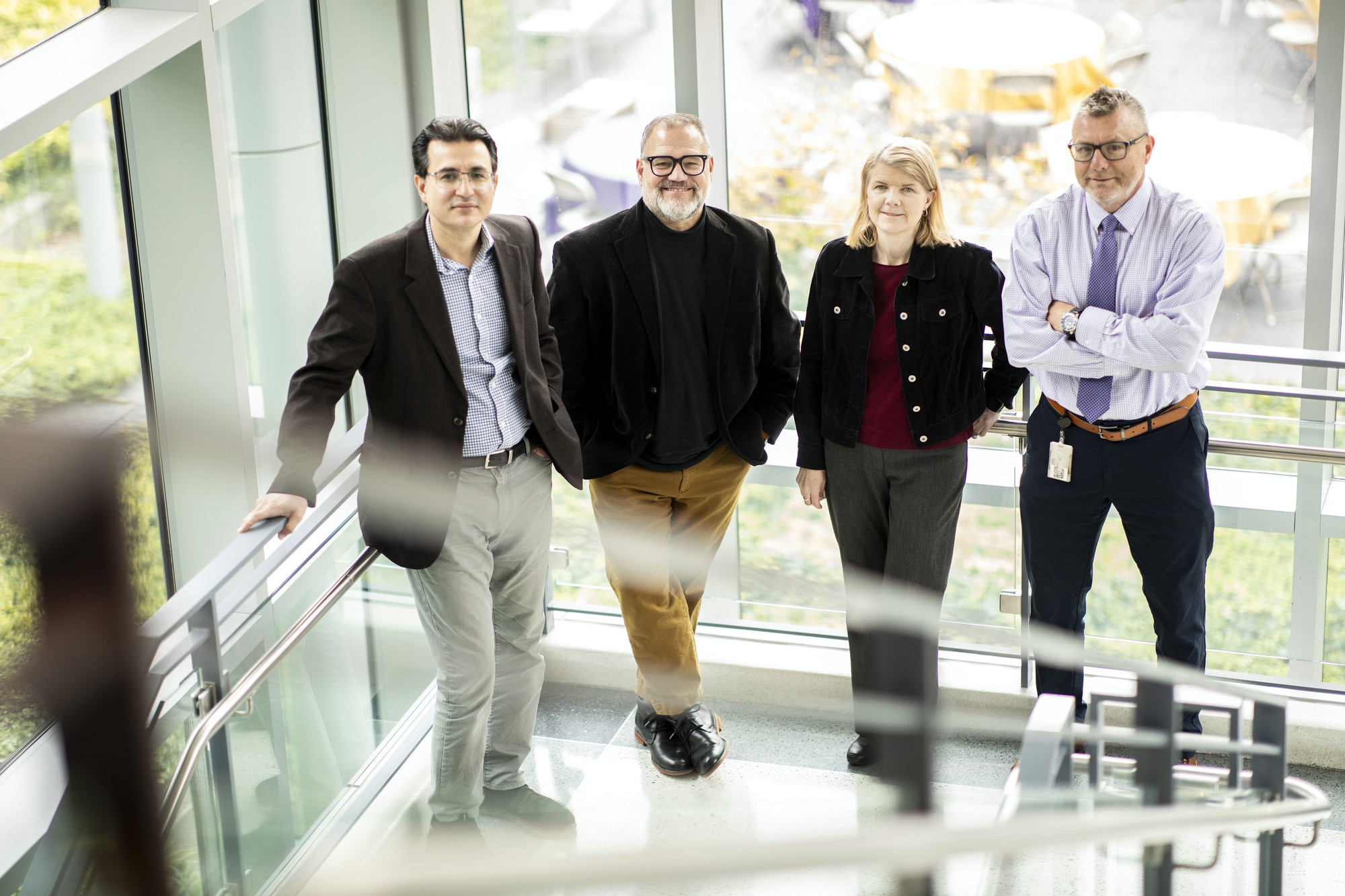 The four featured researchers stand in a light filled glass stairwell in the RNA Institute. From left to right: Mehmet Yigit (man wearing wire rimmed glasses, brown jacket and grey pants), Scott Tenenbaum (man with black glasses wearing black jacket with khaki pants), Melinda Larsen (woman with blond hair wearing a black jacket with gray pants), and Andre Melendez (man wearing black rimmed glasses, pale purple dress shirt and tie and black slacks.