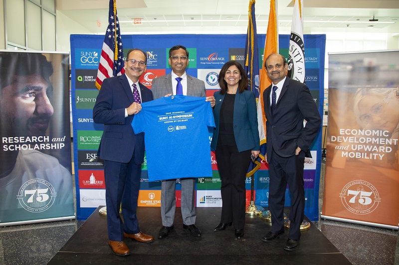 Four people stand in a line in front of U.S., New York State, City of Albany and UAlbany flags smiling and holding a blue SUNY AI Symposium t-shirt.