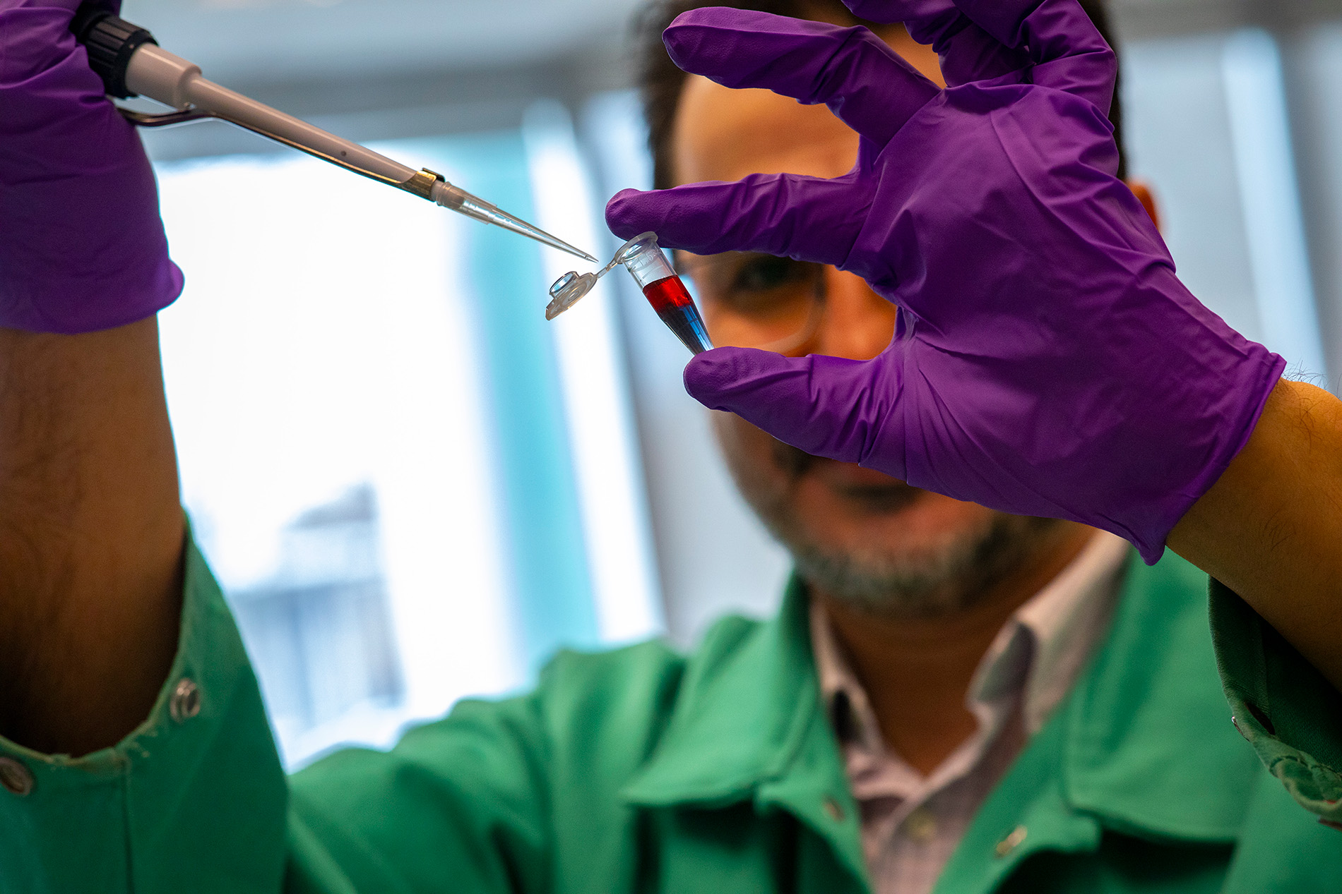 A man wearing a green lab coat and purple gloves holds a vial of solution. The top of the solution is red and the bottom is purple. 