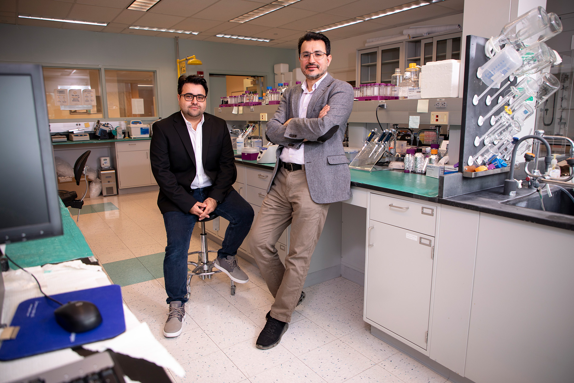 Two men pose for a portrait in a chemistry lab. One is seated, wearing a black blazer and jeans; the other is leaning against a lab bench wearing a gray blazer and khaki pants. Various glass and plastic containers are on the bench and shelf behind them.