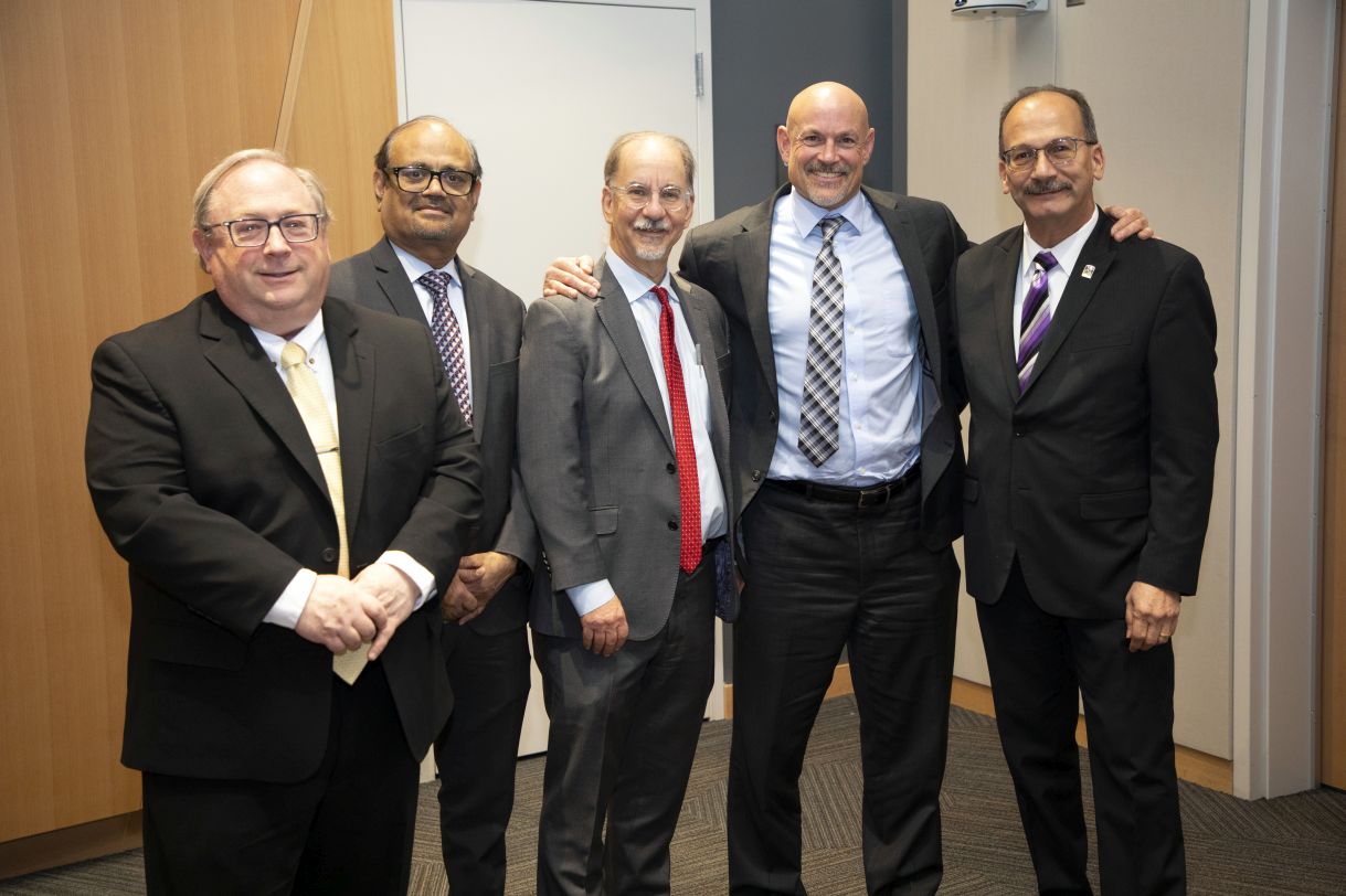 Five men wearing suits pose and smile for a casual group portrait.