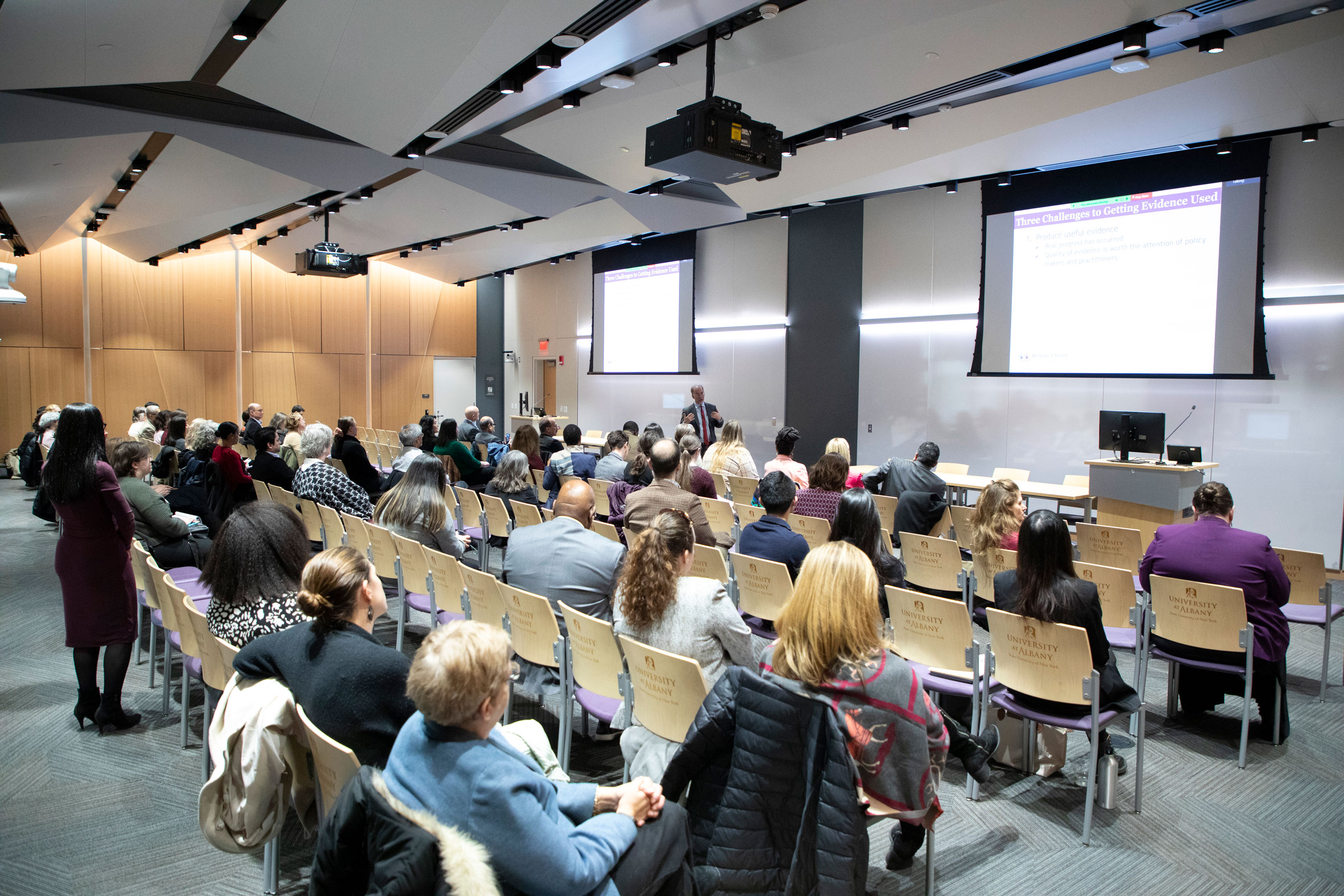 ISHE launch in a room with two projector screens and rows of chairs with people sitting.