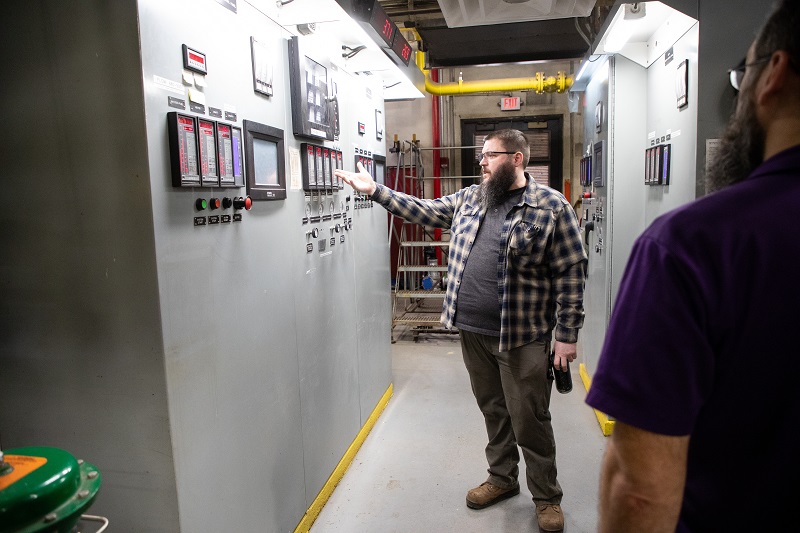 A man stands gesturing to a massive control panel with numerous switches and digital screens.