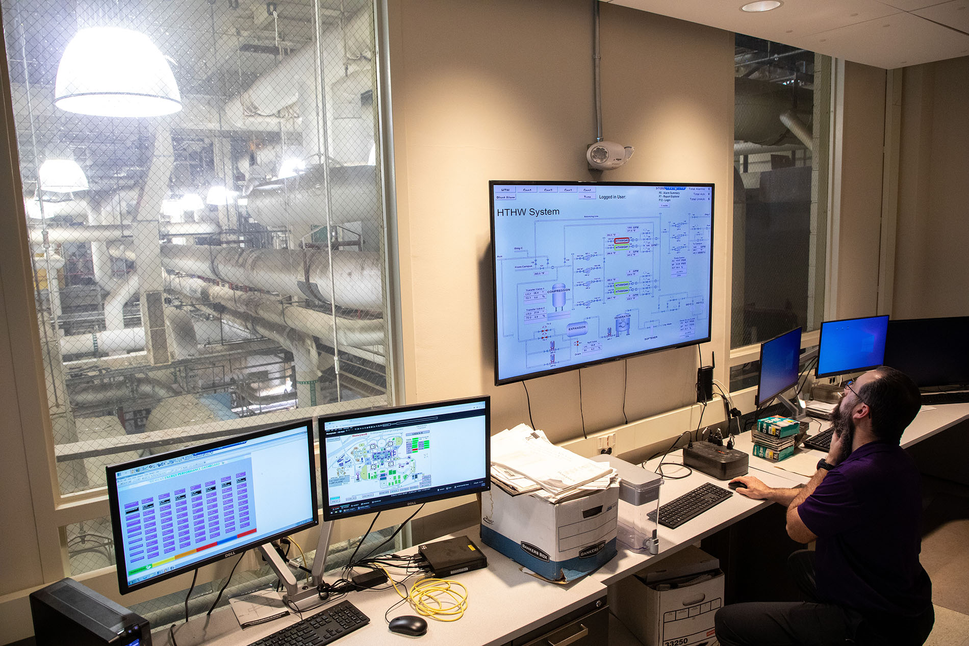 A bearded man sits at a long work station in front of a computer watching several monitors. 
