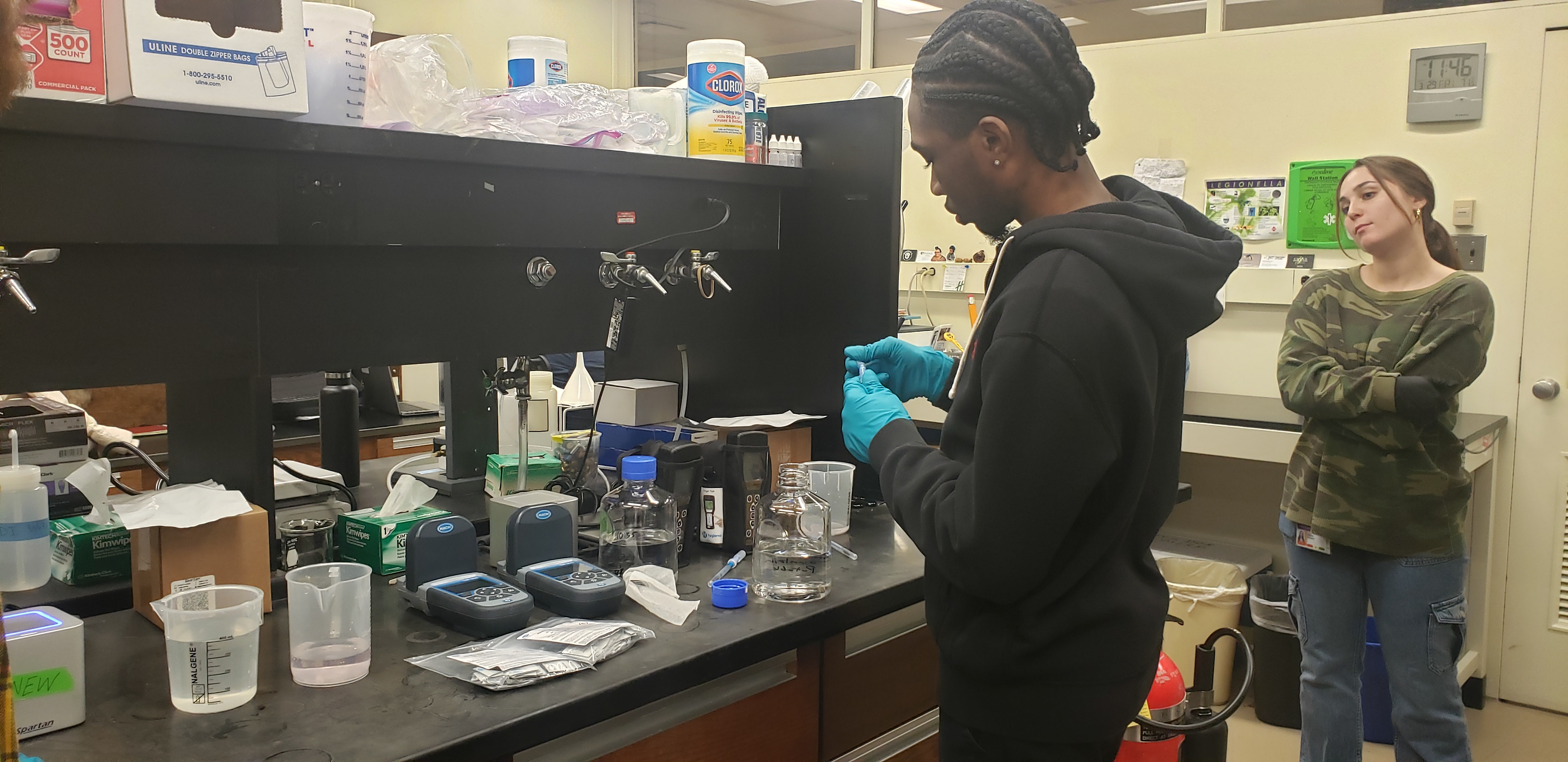 A young man wearing a black hoodie and blue latex gloves stands at a lab bench covered in various instruments used to detect legionella presence. The lab bench is black and the walls in the background are white. A young woman in a camo print shirt looks on. 