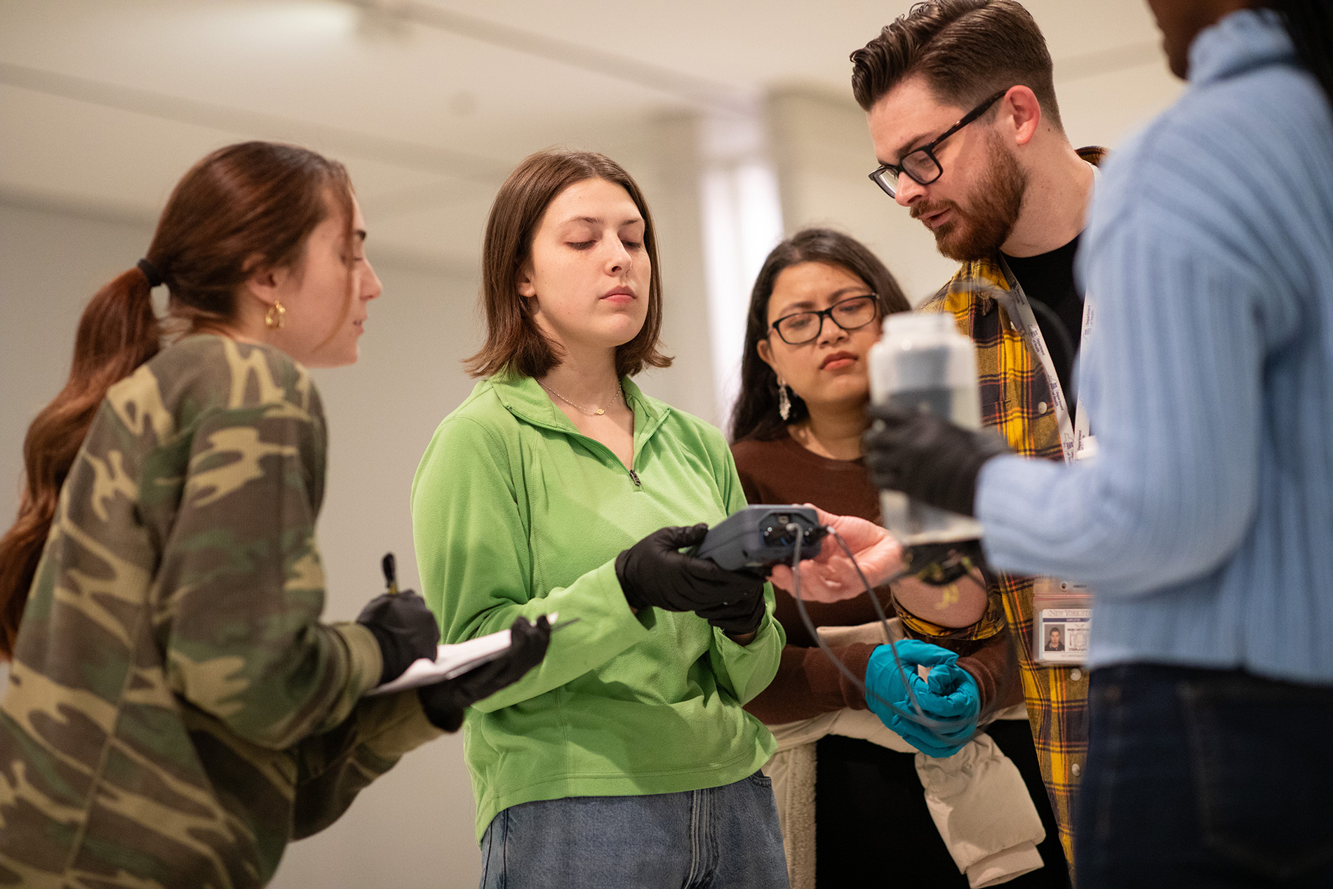 Five young people, four women and one man, stand together holding instruments to collect and record water chemistry measurements. A woman in the center is holding a gray electronic device connected to wires with probes placed in a water bottle held by a woman wearing a blue sweater and black gloves. A woman wearing a camo print shirt holds a clipboard and looks on. A woman in a brown sweater and a man in a yellow plaid shirt also look on. 