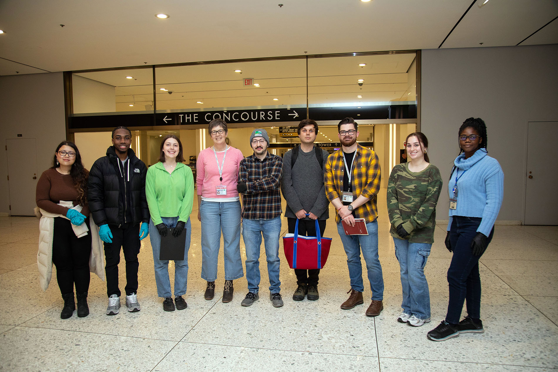 Nine smiling people, five women and four men, stand together for a group portrait under a sign that reads “The Concourse” at Empire State Plaze. Several are wearing latex gloves used to safely collect water samples. – I 