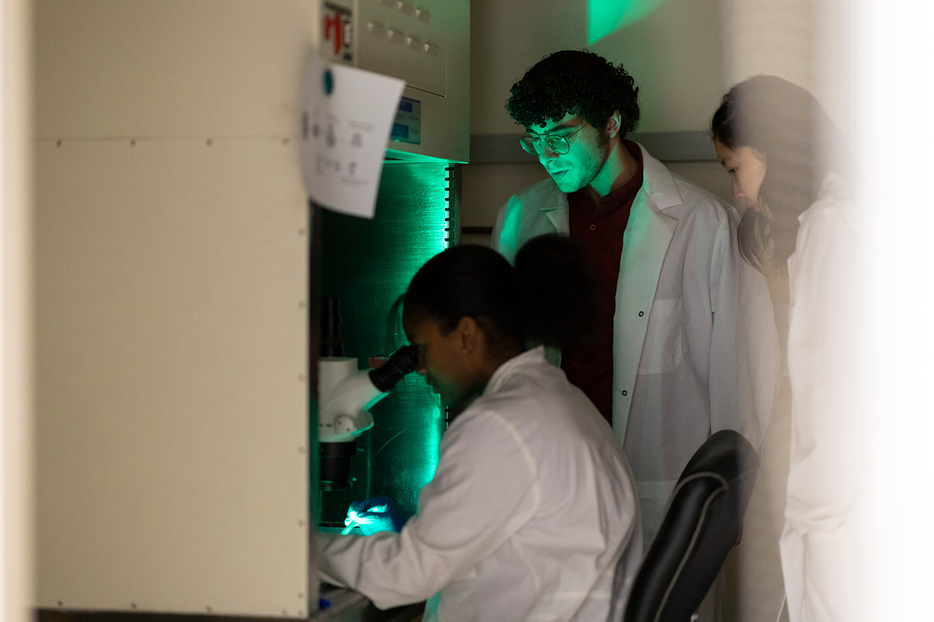 Three students are pictured wearing white lab coats in a darkened room. One is seated, looking into a microscope which is emitting a green glow.