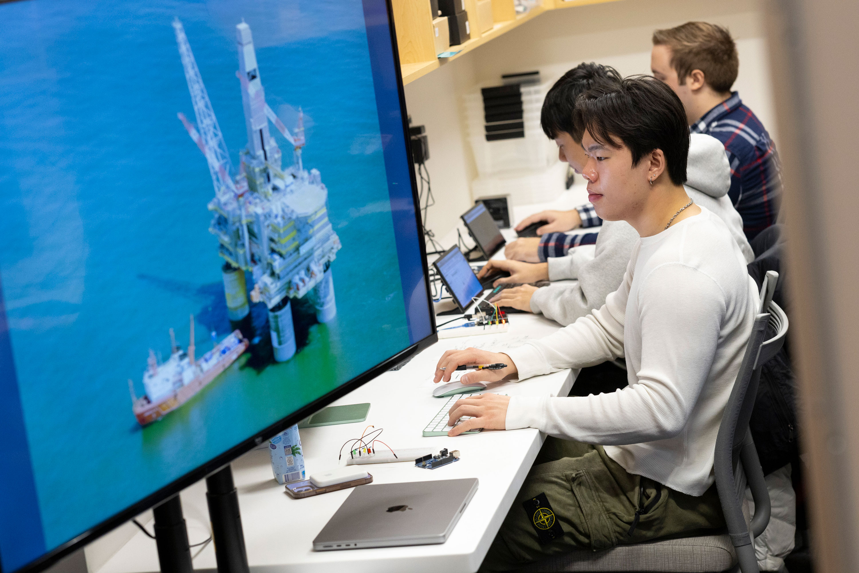 Several students sit at a desk working on laptops and other small devices with a large screen on the side that displays an image of an offshore oil platform surrounded by water.