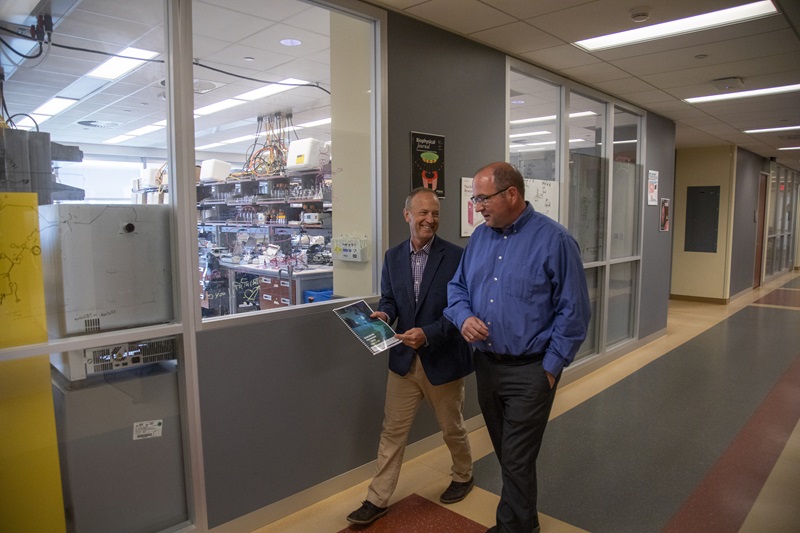 Two men walking down a hallway facing each other in conversation while one gestures to the papers in his hands.