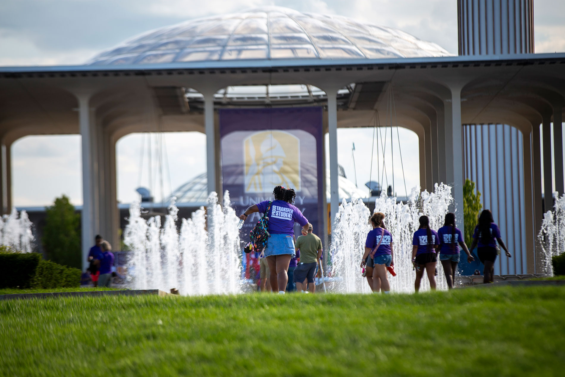 Students walking through the entrance fountain wearing Proud Transfer shirts.