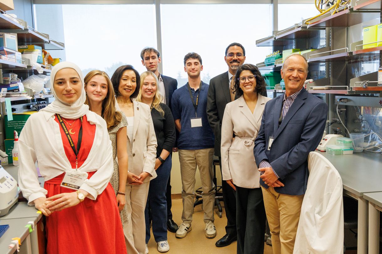 Nine smiling people stand together in a lab, posing for a group portrait. They are flanked by lab benches and shelving stocked with lab supplies.