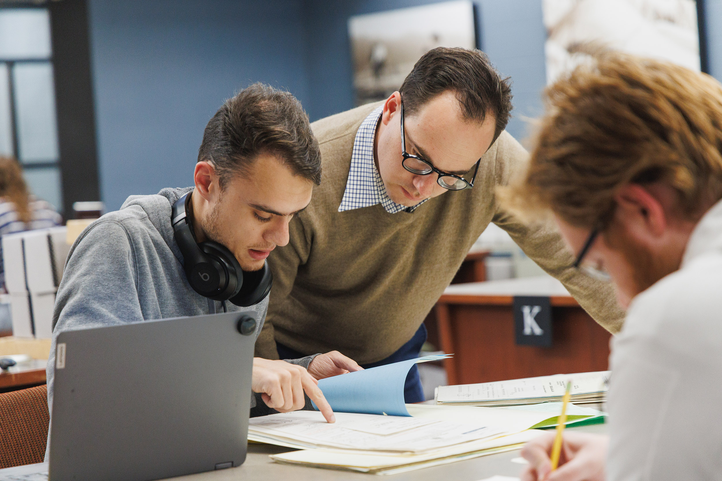 A man in a gray sweatshirt is seated at a table reviewing a folder of papers as a man in a brown sweater with glasses looks over the documents with him.