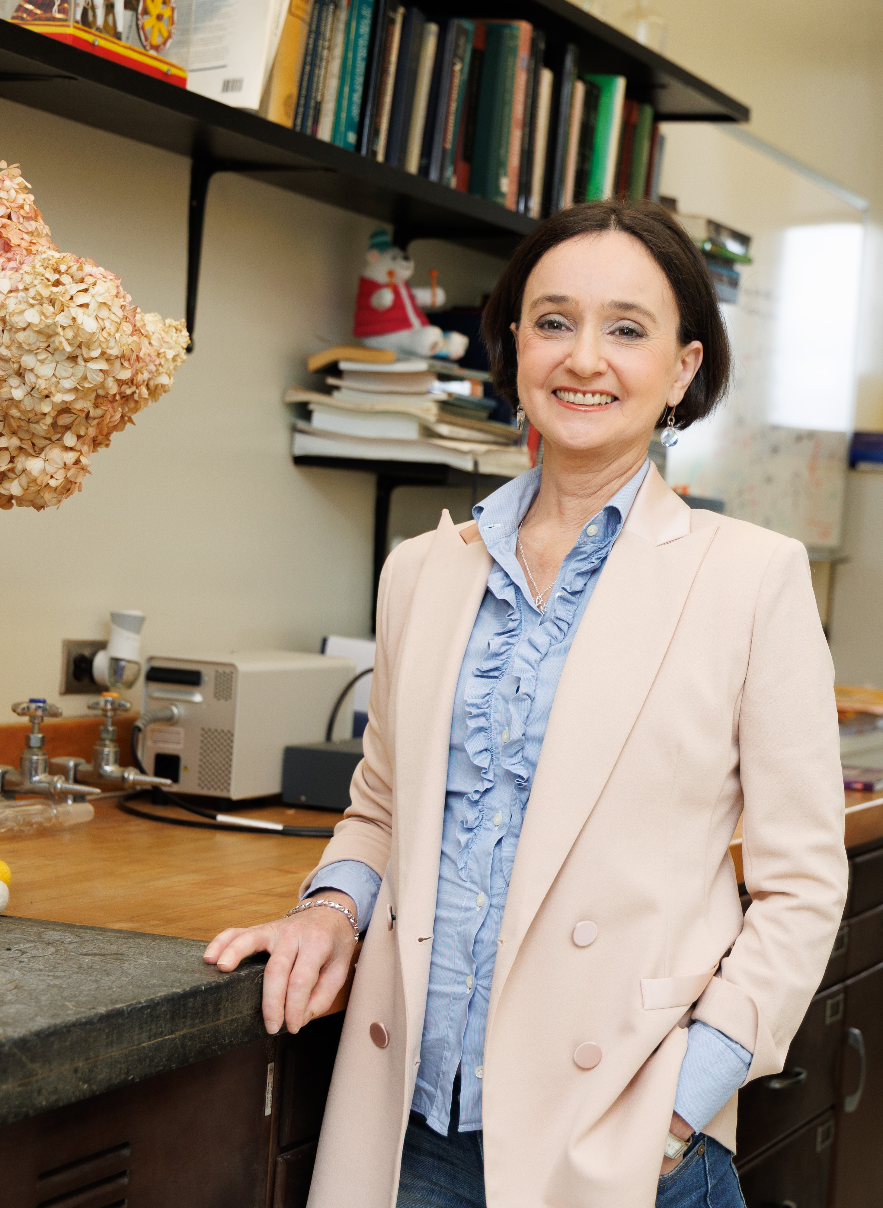A woman with short brown hair smiles for a portrait. She is wearing a pale pink blazer with a frilled, pale blue blouse underneath.