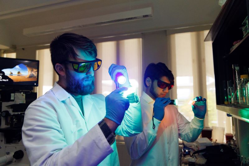Two me in lab coats and protective glasses hold up microscope slides illuminated by flashlights casting a blue-green light.