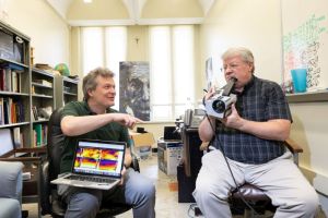 Two men seated facing the camera; one has a laptop on his lap showing a screen with bright colors, while the other is holding a gray camera.