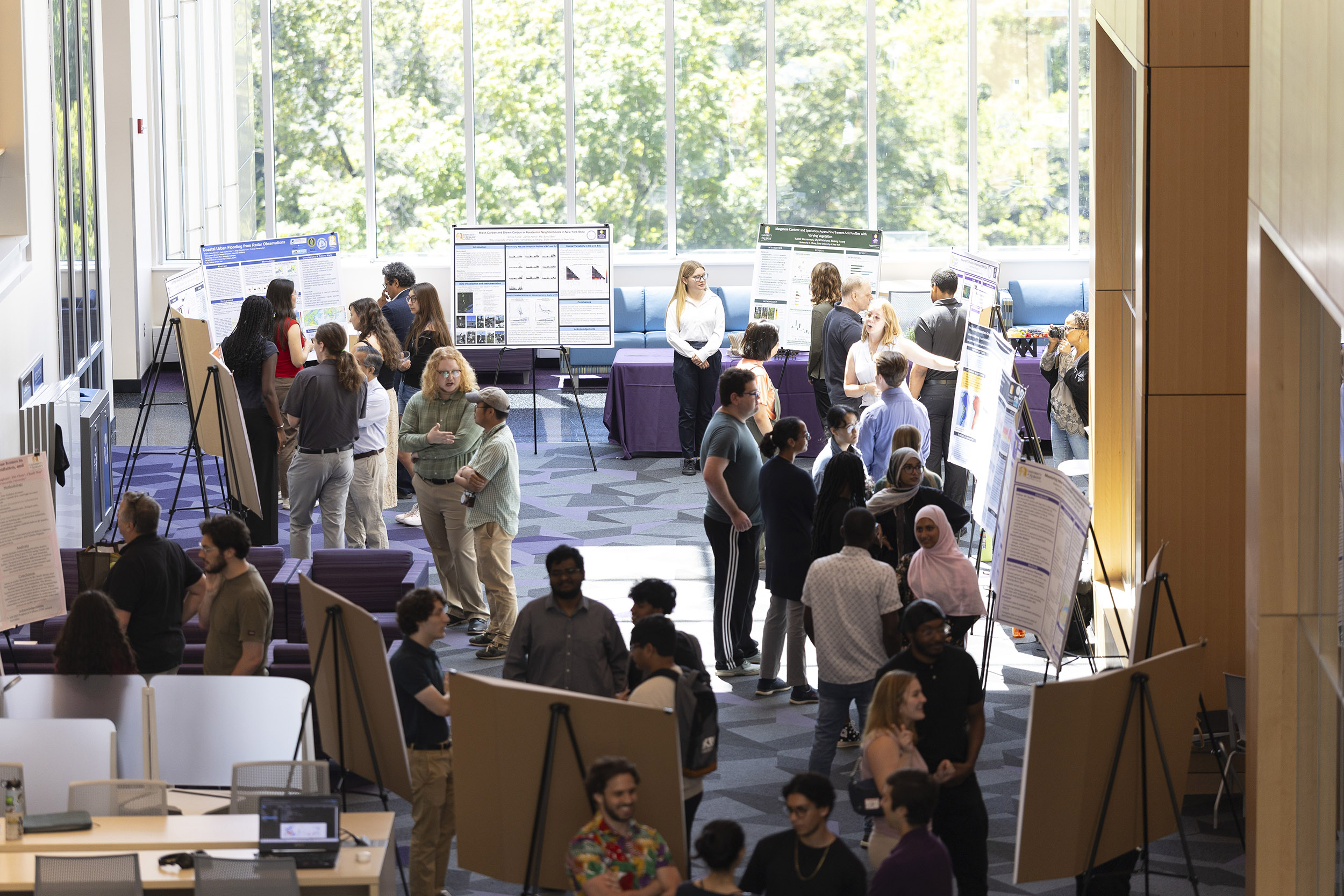 Several students stand in front of numerous posters in the foyer of an acadmic building in front of a row of glass windows along the back wall.