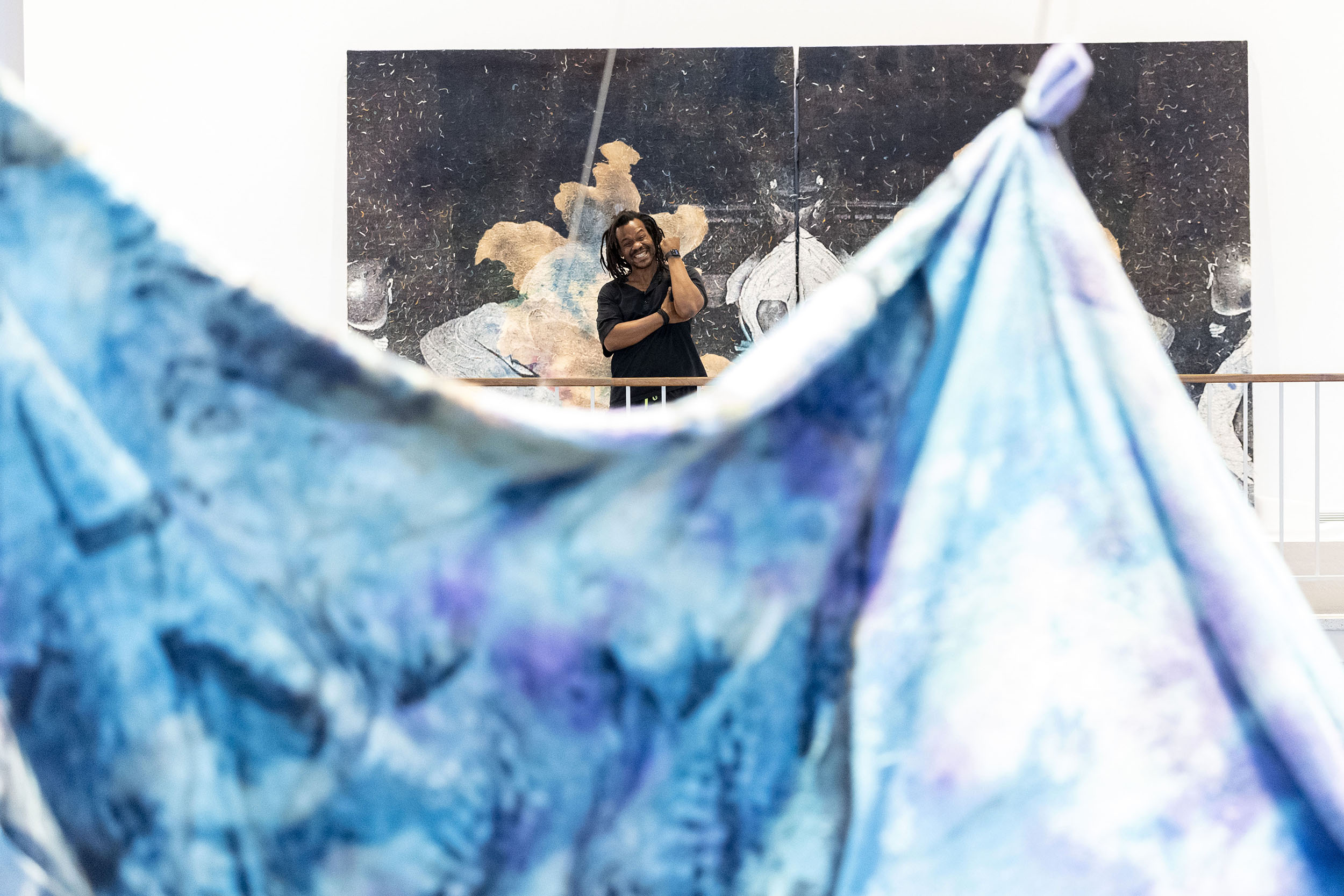 An African American man stands in the background as a colorful tapestry is suspended in front of him in a room with white walls.