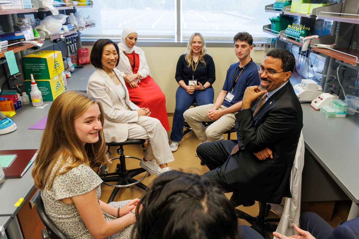 Seven smiling people are seated in a circle in a lab having a conversation.