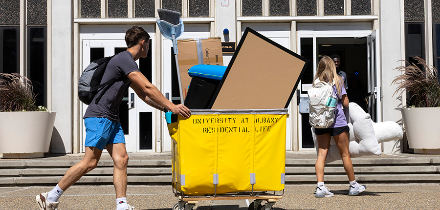 Students wheel a bin of items on-campus. A frame, broom and pillow can be seen in the bin.
