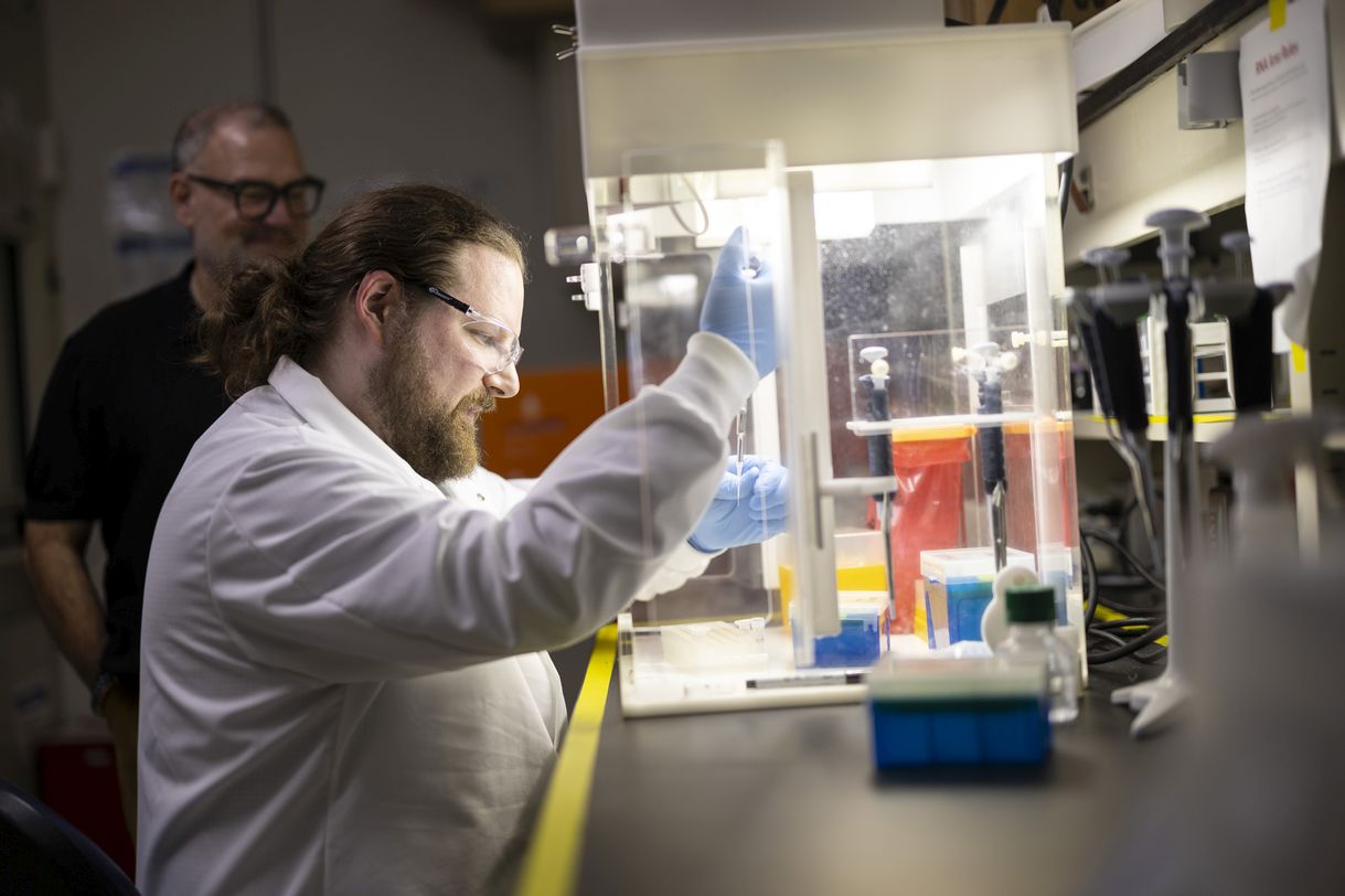 A man wearing a white lab coat, blue latex gloves and clear protective glasses handles a pipette as a man with black rimmed glasses looks on in the background.  