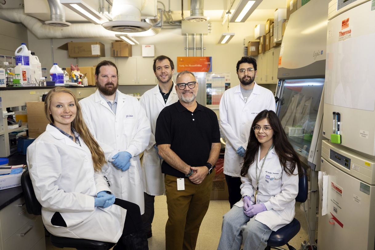 Six people pose for a group portrait in a brightly lit lab. Five are wearing white lab coats; the man in the center is wearing square black rimmed glasses and a black polo shirt. 