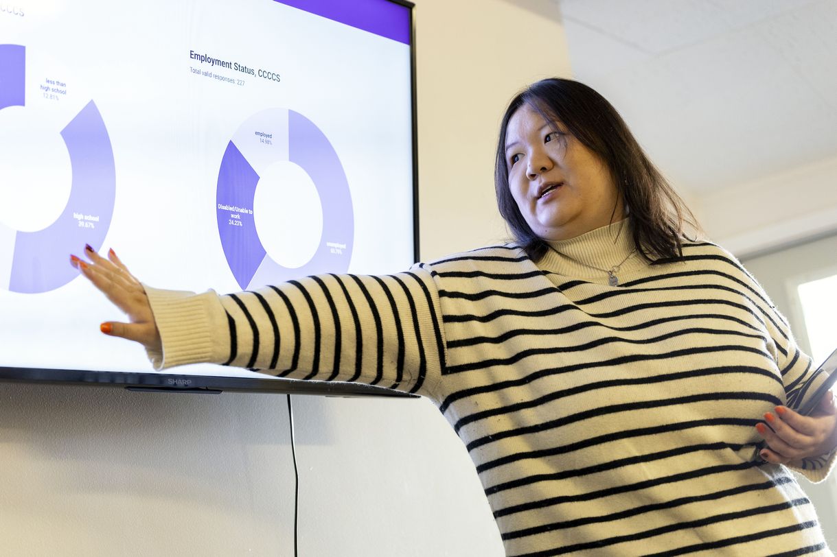 A young woman with short brown hair wearing a white and black striped sweater and holding an iPad gestures to a glowing PowerPoint screen displaying purple pie charts.