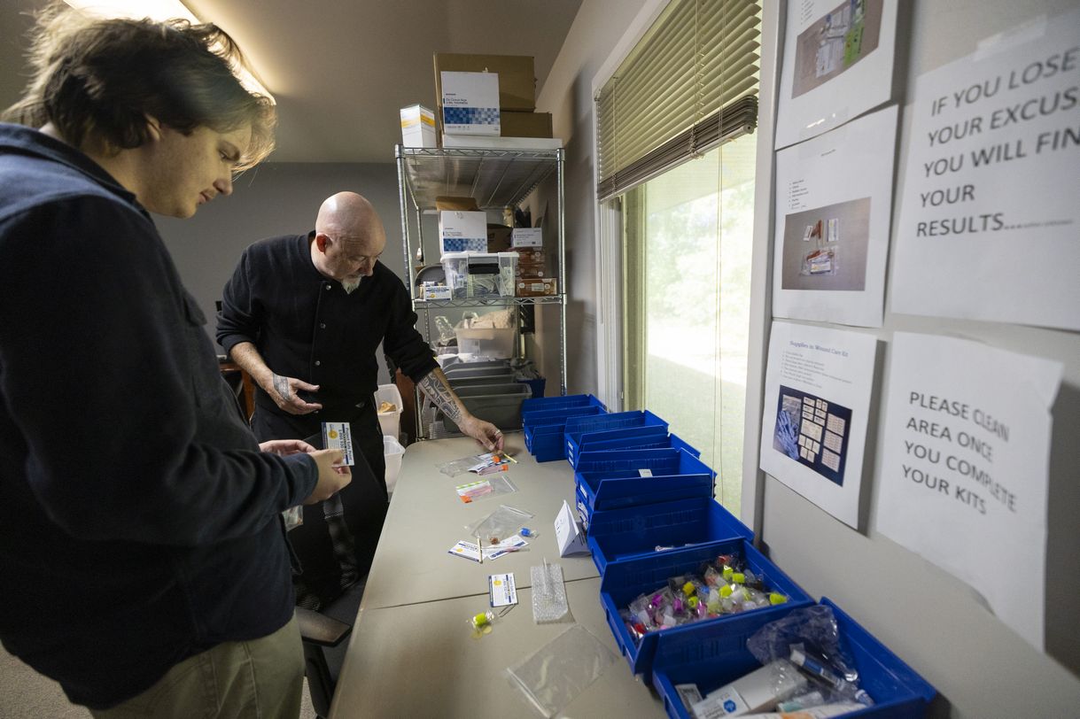 Two men stand in a room containing shelves laden with supplies. They are packing small clear plastic bags with various 'safer smoking' implements carefully organized into blue plastic bins.