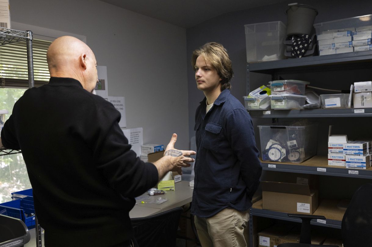 Two men stand in a gray-painted room containing shelves stocked with various care and medical supplies.
