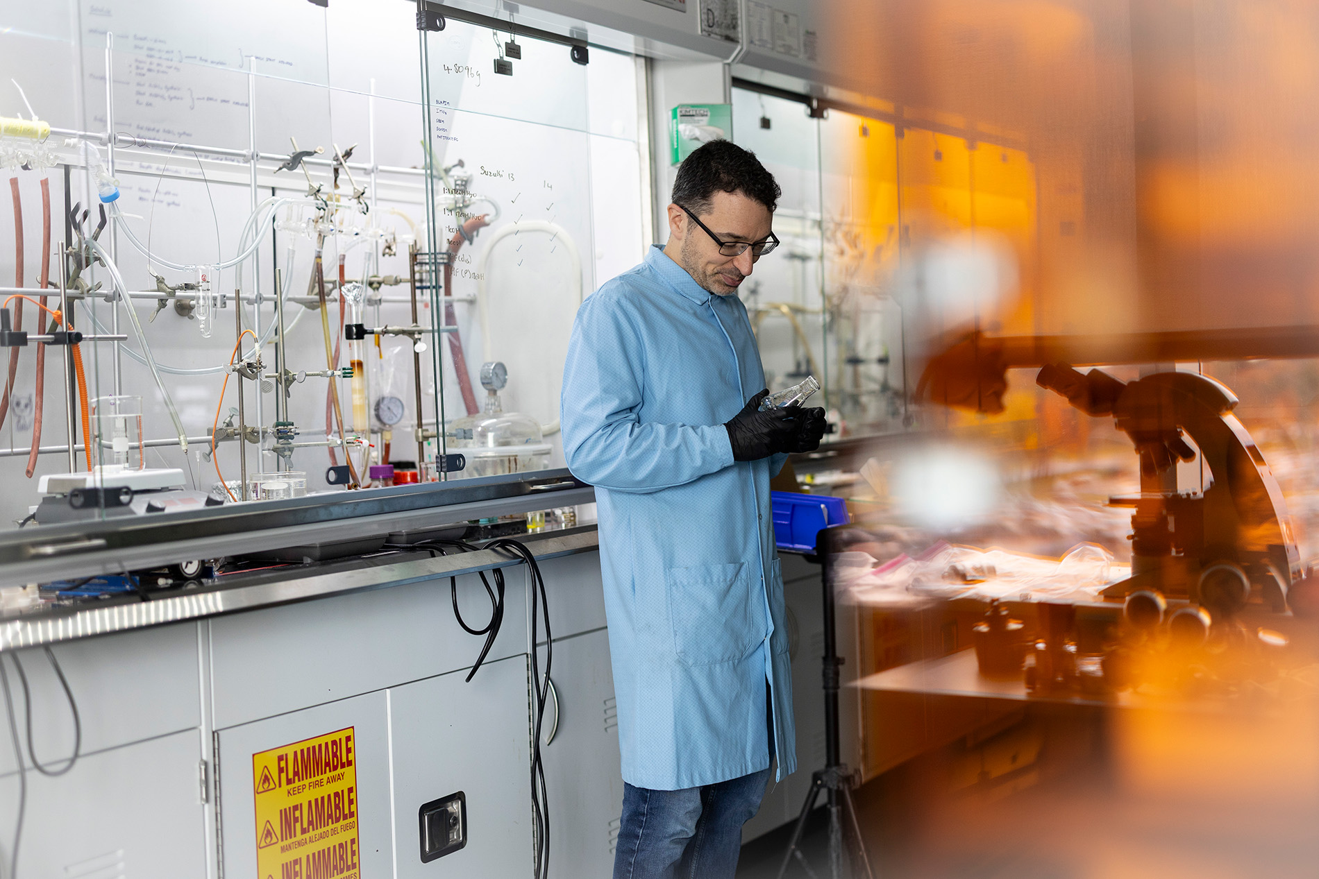 A man wearing a pale blue lab coat and black gloves examines a beaker in a chemistrylab.