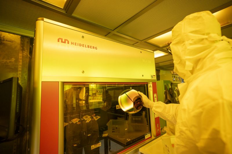 A person in an all white cleanroom suit holds up a silicon wafer in front of a large piece of lab equipment.