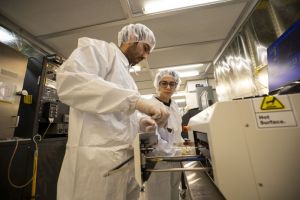 Two people in white cleanroom suits insert a silicon wafer into the drawer of a white lab tool.