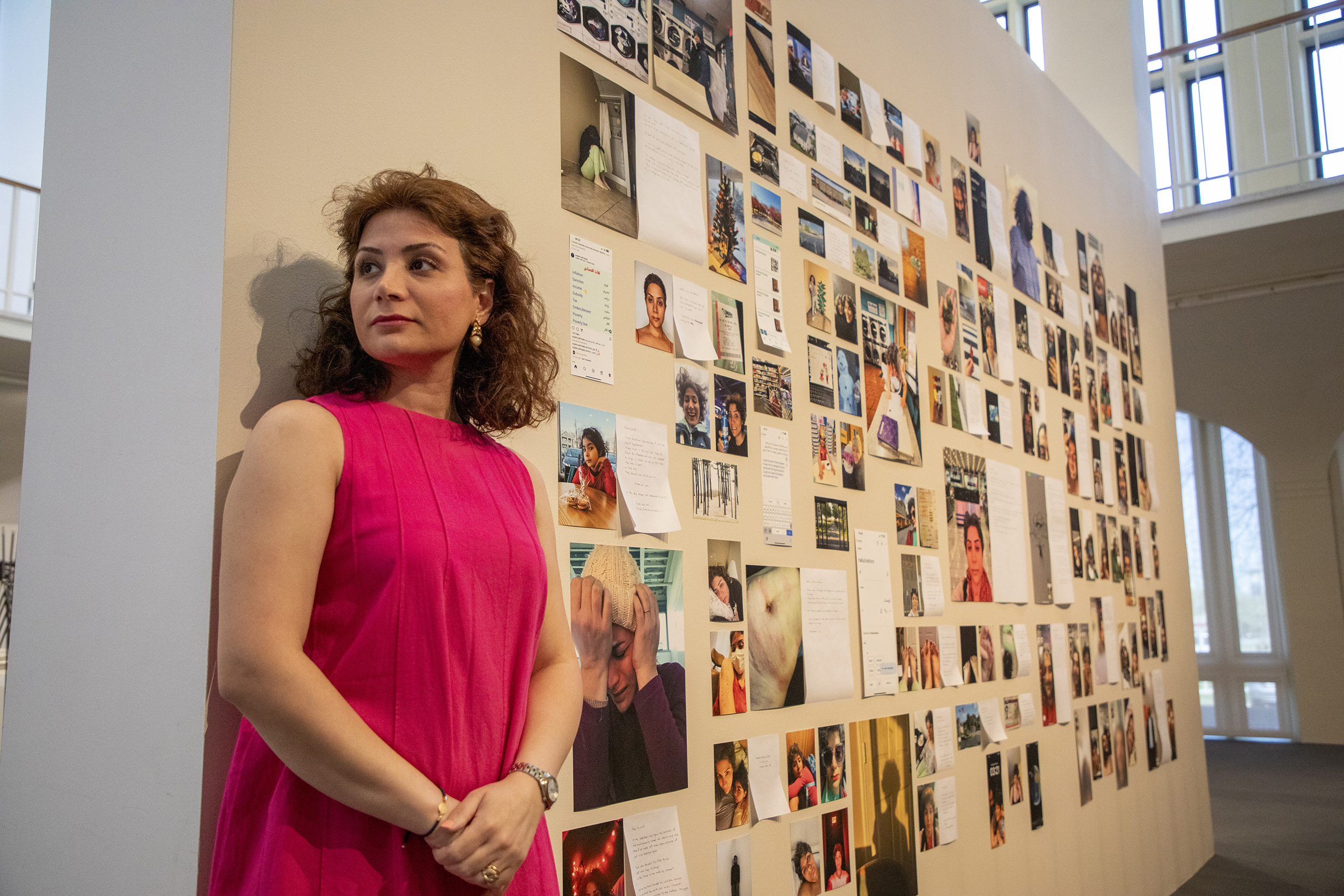 A woman with brown shoulder-length hair, earrings in a bright pink blouse stands along a wall looking into the distance as many photos are on display along the wall behind her.