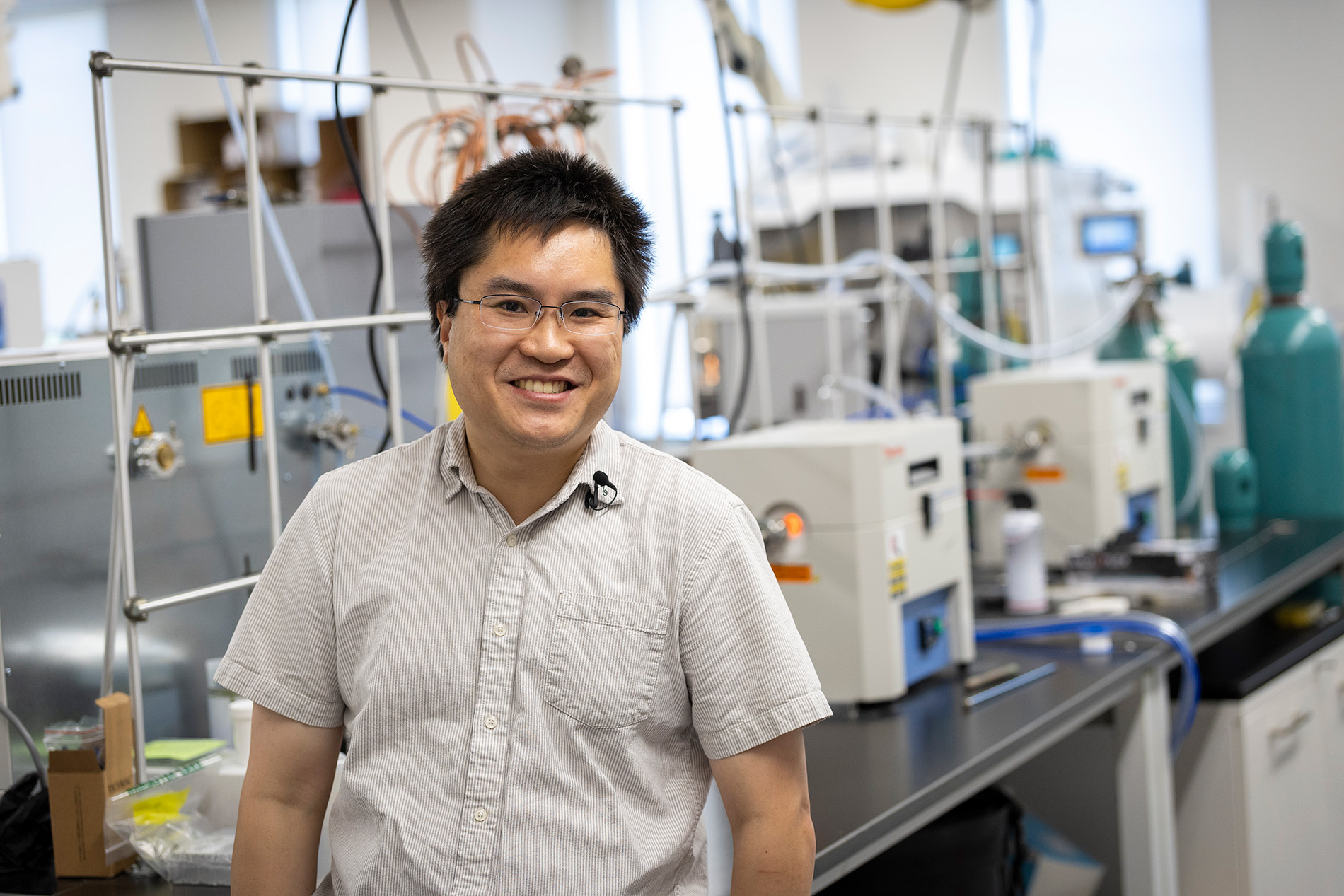 A man wearing a short sleeved button down shirt and glasses stands in front of a lab bench, smiling. 