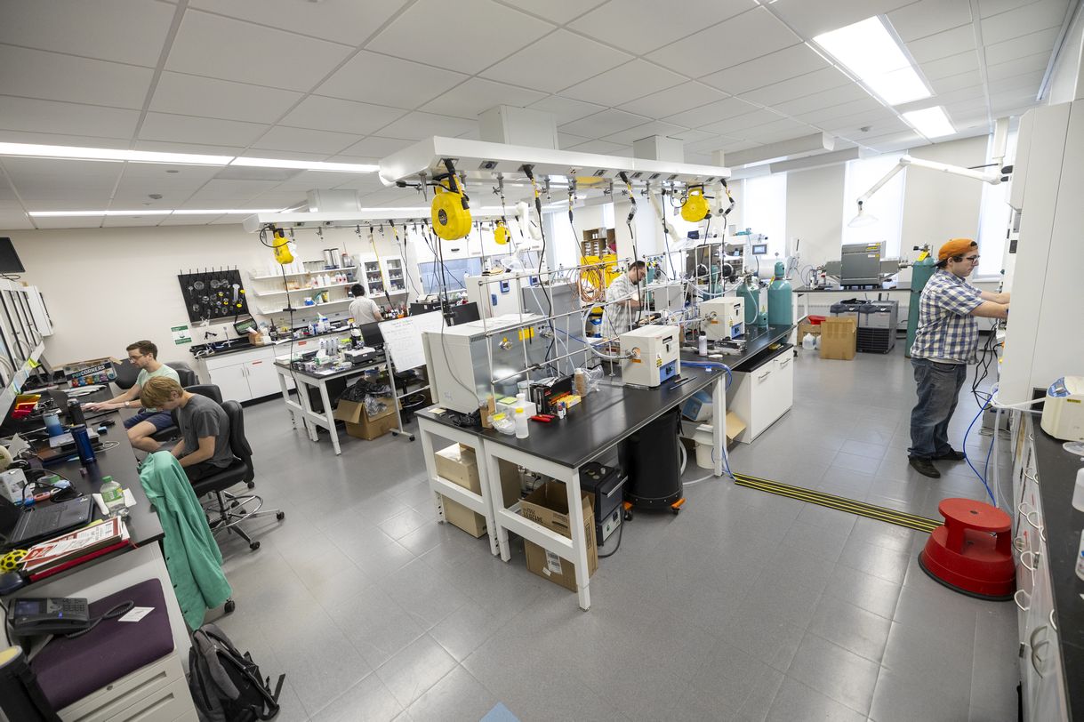  Image shows a brightly lit lab full of black-topped lab benches stocked with various pieces of equipment connected to cords hanging from the ceiling.