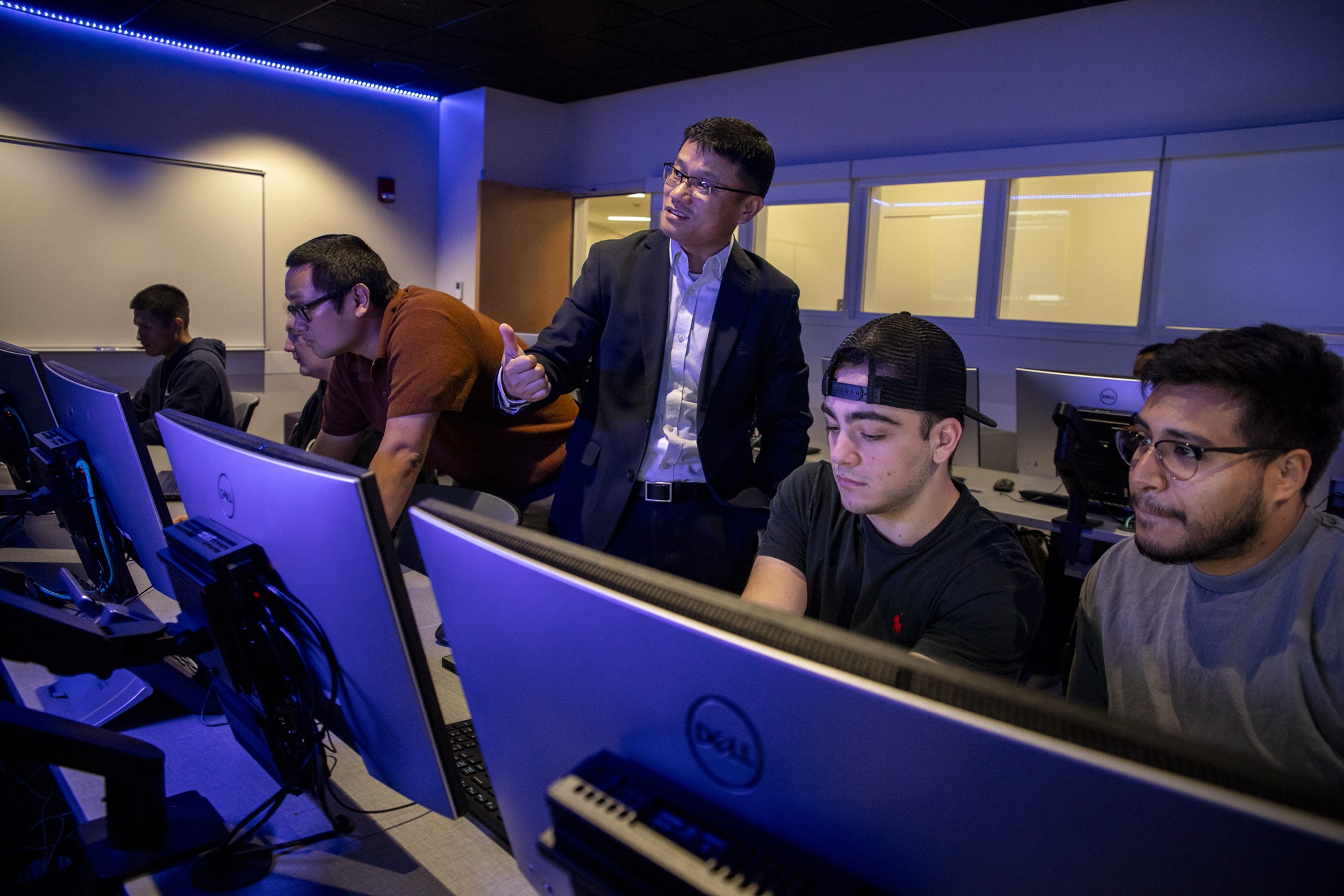 Alan Wang stands between students reviewing code at the Cyber Range lab.