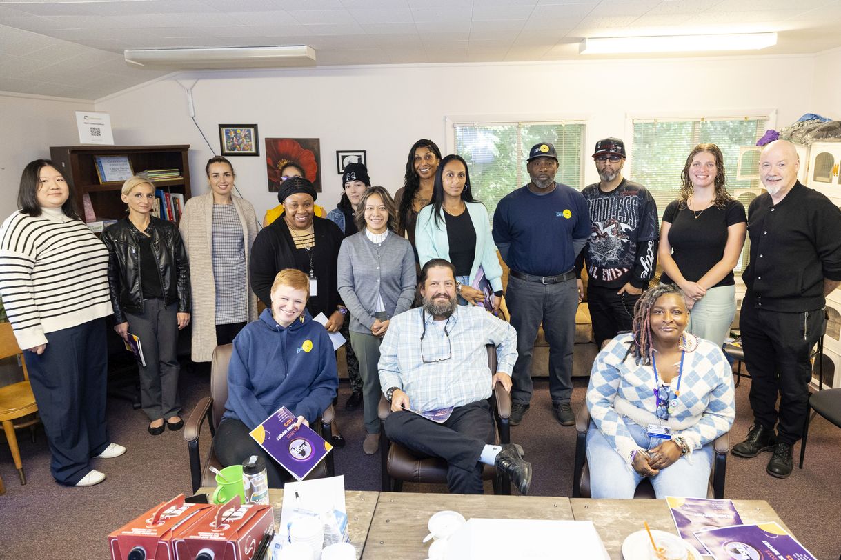 A group of sixteen people pose for a group portrait in a common area at Project Safe Point's Albany location. The room has multiple windows, artwork hung on the walls, and is lined in shelves containing books and bins of personal care supplies.