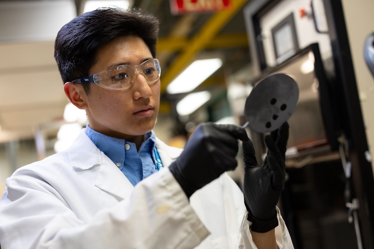 A man with short black hair in safety goggles, white lab coat and black gloves inspects a piece of lab equipment.