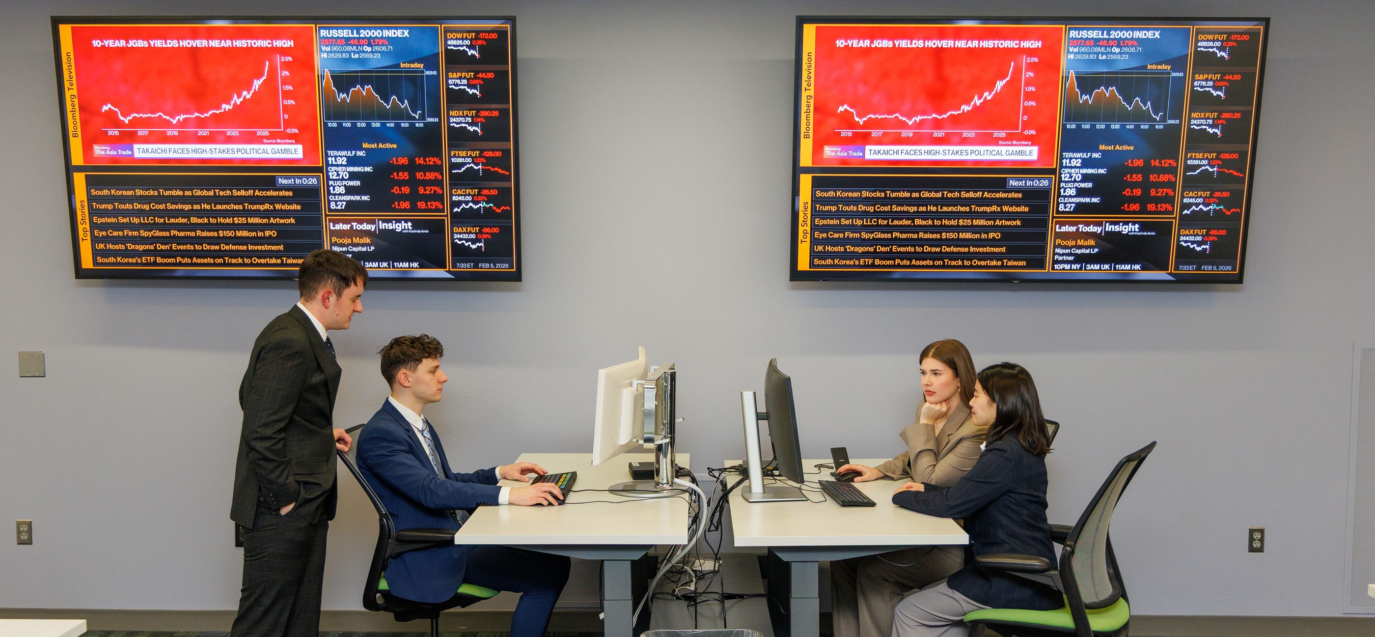 Three UASBIG Group students are sitting at two desks looking at computers. One student is standing behind another student that is sitting. On the wall behind them are two televisions with research.