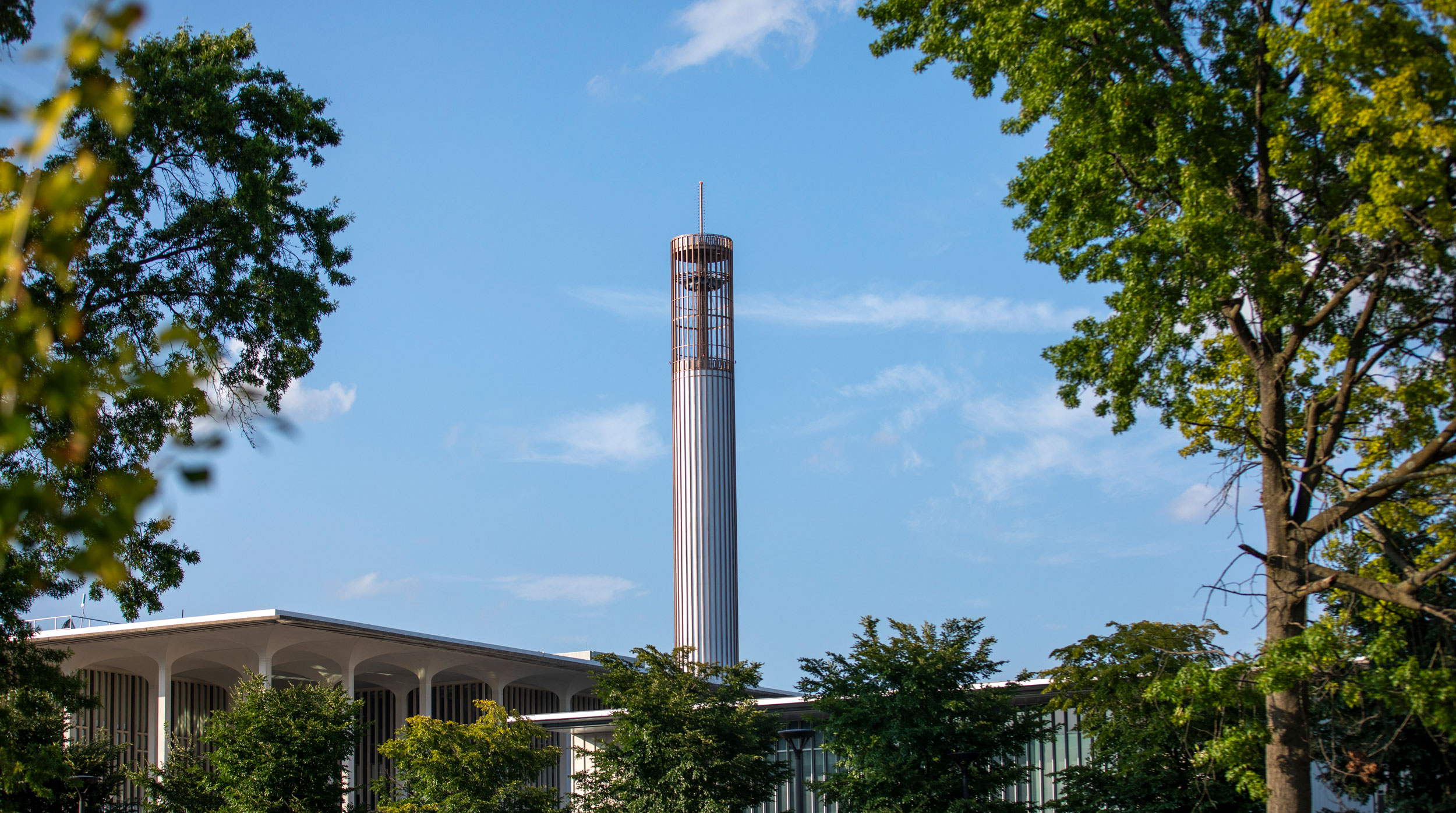 The UAlbany carillon, framed by lush green trees and photographed against a bright blue sky.