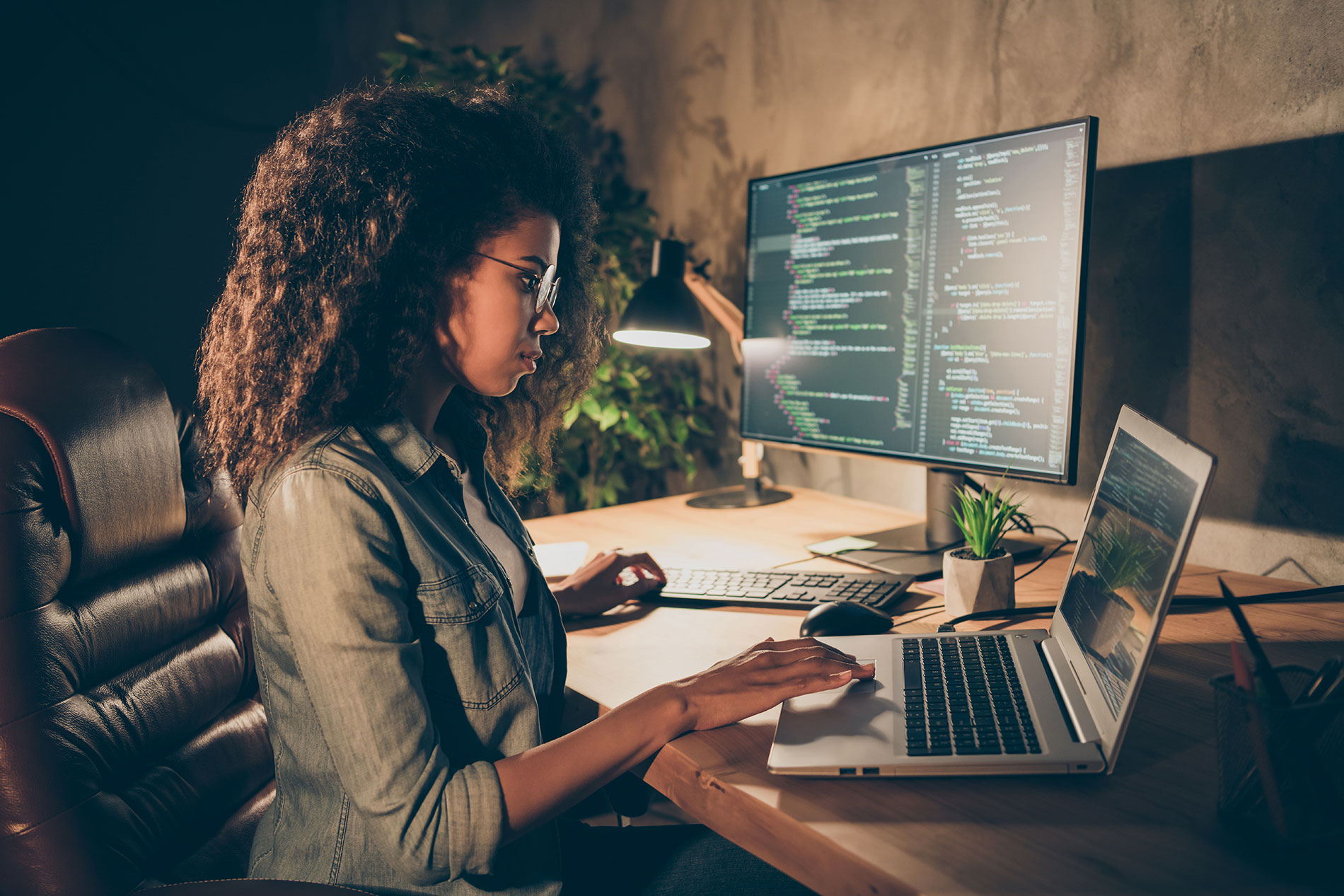 A cybersecurity professional works on a laptop alongside a monitor displaying code.