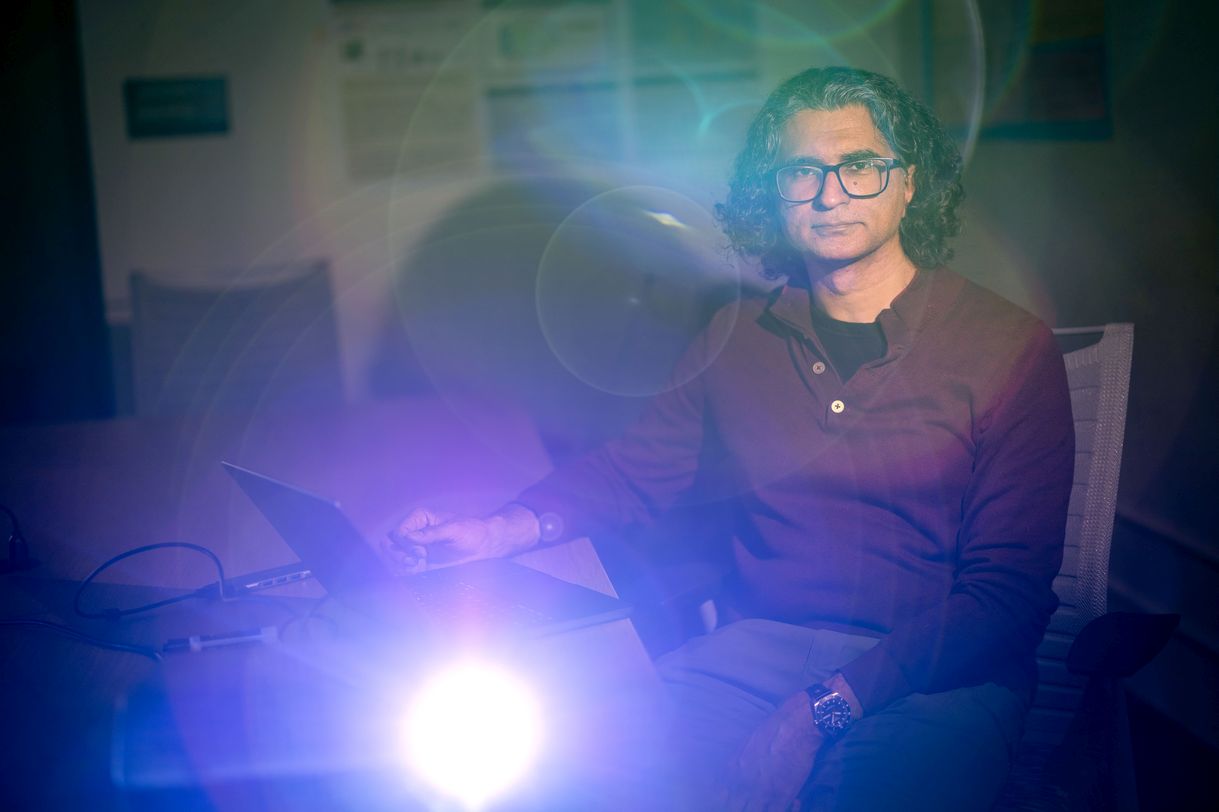 A man wearing a maroon shirt and glasses sits in a dark classroom lit by a projector creating ethereal orbs of light across the image.