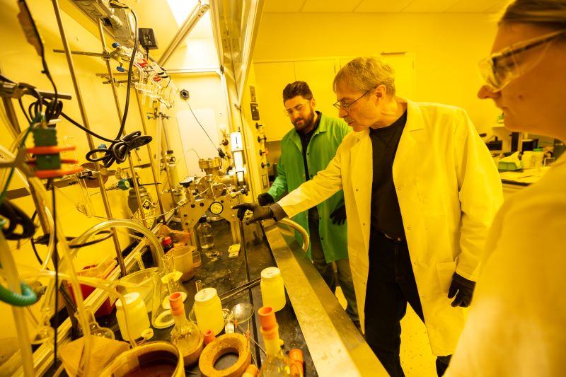 Three people in lab coats stand at a lab bench looking at a piece of equipment in yellow-lit chemistry lab.