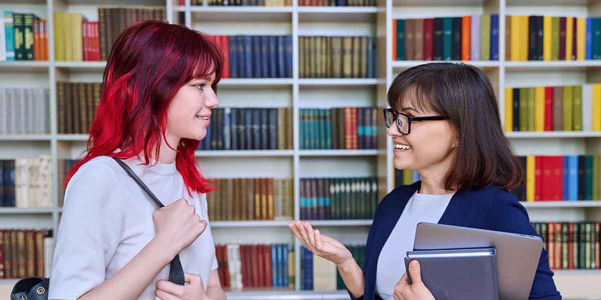 An academic advisor talks to a student in the school library.