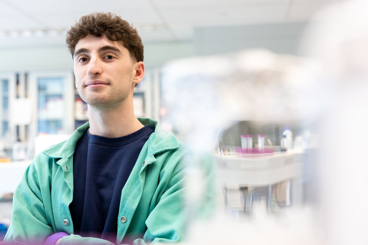 A young man with dark hair wearing a green lab coat stands in a brightly lit lab. The background is artfully blurred. 